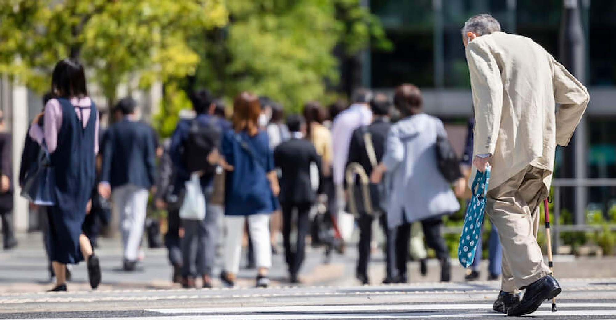 写真：横断歩道を歩く人々と、遅れて辛そうに歩く老人